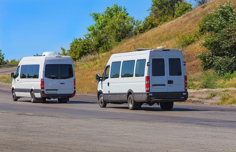 Two Passgenger vans drive on a road with trees in the background