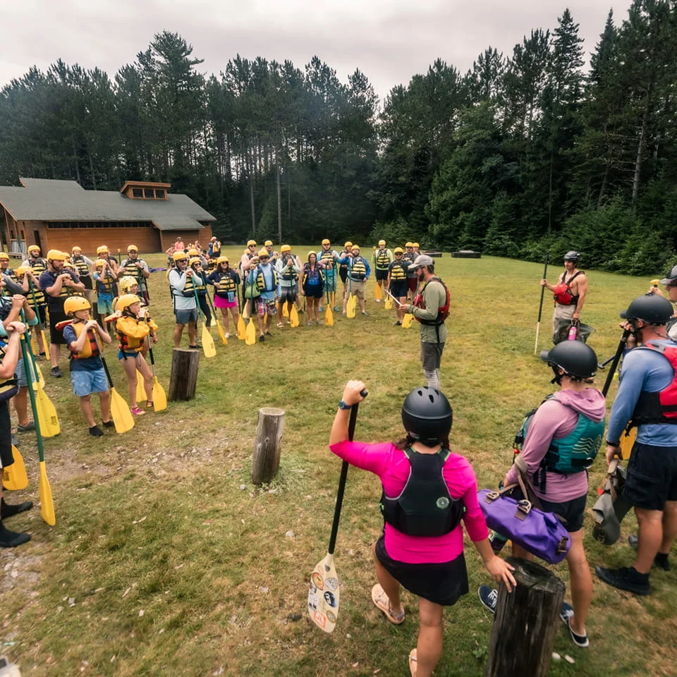 group of people wearing helmets and lifejackets, and holding paddles, listening to a safety briefing from their rafting guides